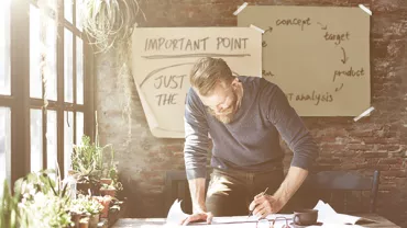 Man werkt aan bureau met papieren en kamerplanten, brainstormende aantekeningen op muur.