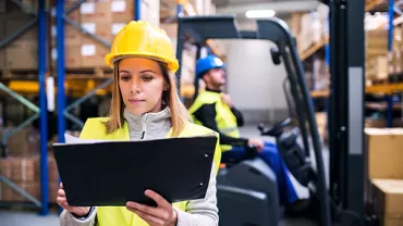 Vrouw in magazijn controleert clipboard; heftruck op de achtergrond.