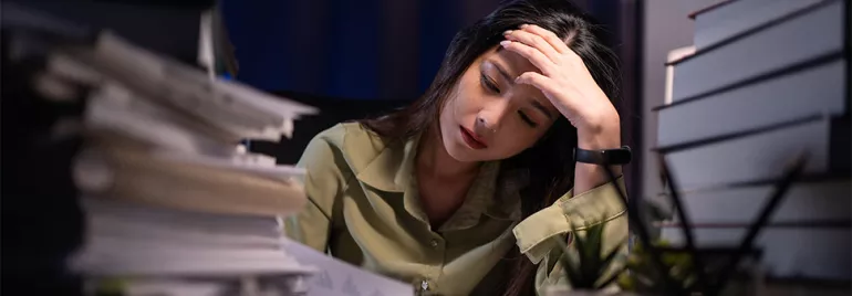 Stressed woman in office, facing work stress and burnout, surrounded by documents at her desk.